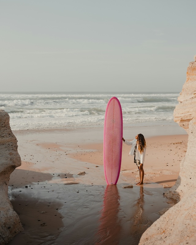 Looking through rocks at a beach with the sea behind. The sand is wet and shiny. A woman stands on it 
        holding a long paddle board.